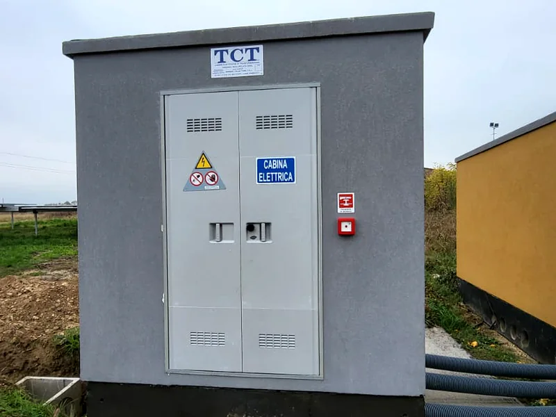 Cabina prefabbricata TCT grigia posata su una platea, accanto a un impianto fotovoltaico