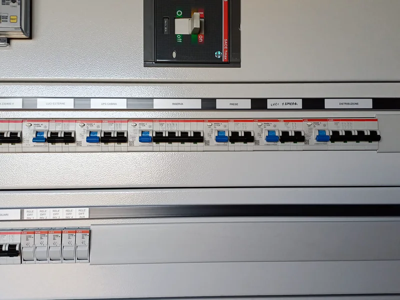 Fila di interruttori del quadro dei servizi ausiliari, con le etichette: luci, prese, UPS