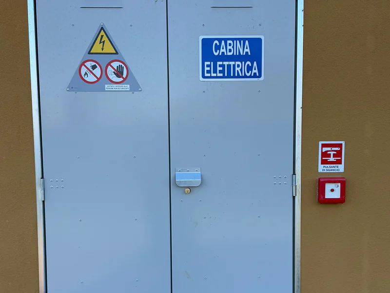 Porta doppia di una cabina elettrica coi cartelli di sicurezza e il pulsante di sgancio accanto